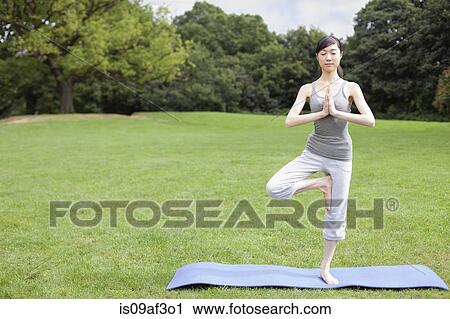 Stock Image - Young woman in park practicing yoga tree pose. Fotosearch