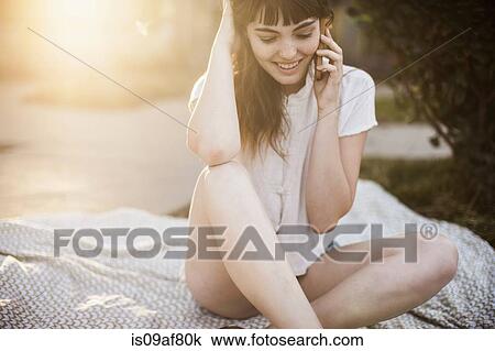 Stock Image - Young woman sitting on picnic blanket talking on cellphone. Fotosearch