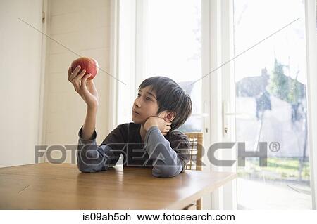 Stock Image - Boy sitting at table, holding an apple in front of him. Fotosearch