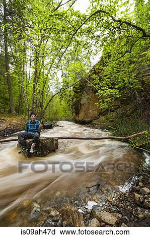 Stock Image - Male hiker sitting on flooded river rock, Naramata, British Columbia, Canada. Fotosearch