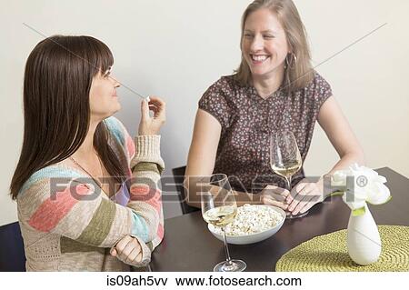 Stock Photo - Mid adult female friends drinking white wine and eating popcorn. Fotosearch
