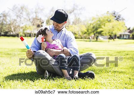 Stock Photo - Mid adult man and daughter eating ice lollies in park. Fotosearch