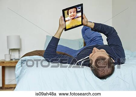 Stock Image - Mid adult man lying on bed holding up digital tablet. Fotosearch