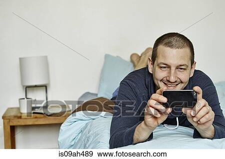 Stock Photo - Mid adult man lying on bed taking self portrait on cellphone. Fotosearch