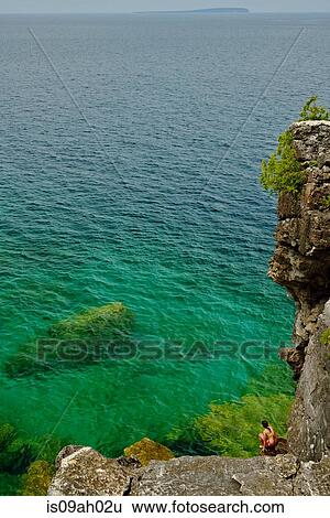 Stock Photograph - Mid adult woman crouching on rocks at coast, Toronto, Ontario, Canada. Fotosearch