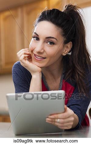 Stock Image - Portrait of young woman in kitchen holding digital tablet. Fotosearch