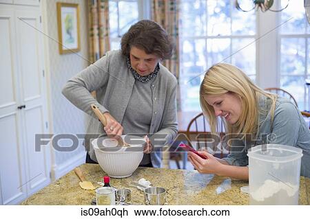 Stock Image - Senior woman and granddaughter baking and using cellphone. Fotosearch