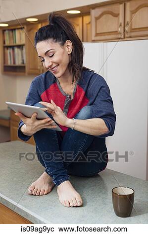 Stock Image - Young woman in kitchen using digital tablet. Fotosearch