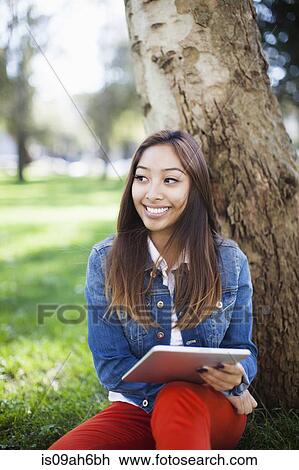 Young woman leaning against tree, using digital tablet View Large Photo Image Stock Image - Young woman leaning against tree, using digital tablet. Fotosearch