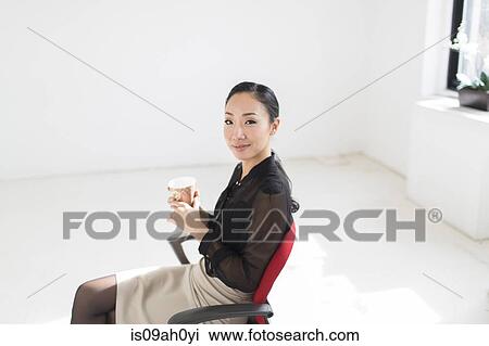 Stock Image - Young woman sitting on office chair with coffee. Fotosearch