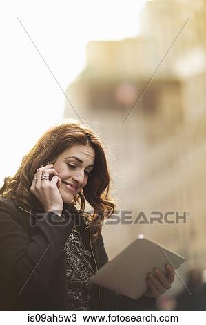 Stock Image - Young woman using cellphone and digital tablet on street. Fotosearch