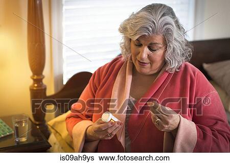 Stock Photograph - Mature woman sitting on bed reading information on pill bottle. Fotosearch