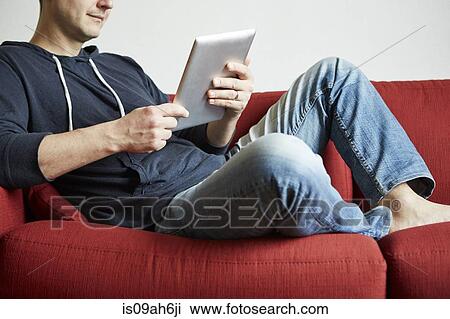 Mid adult man relaxing on sofa looking at digital tablet View Large Photo Image Stock Image - Mid adult man relaxing on sofa looking at digital tablet. Fotosearch
