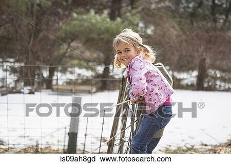 Stock Image - Young girl climbing wire fence in field. Fotosearch