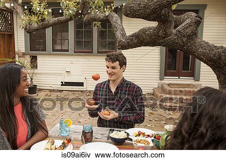 Friends sitting around table sharing barbecue food, man juggling fruit View Large Photo Image Stock Photo - Friends sitting around table sharing barbecue food, man juggling fruit. Fotosearch