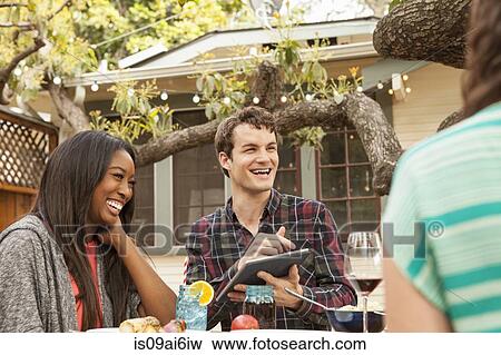 Friends sitting around table sharing barbecue food, man using digital tablet View Large Photo Image Stock Photo - Friends sitting around table sharing barbecue food, man using digital tablet. Fotosearch