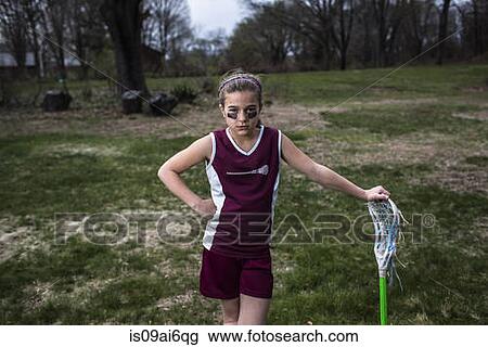 Stock Image - Girl wearing lacrosse uniform, leaning against lacrosse stick. Fotosearch
