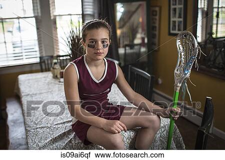 Stock Image - Girl wearing lacrosse uniform, sitting on dining table. Fotosearch