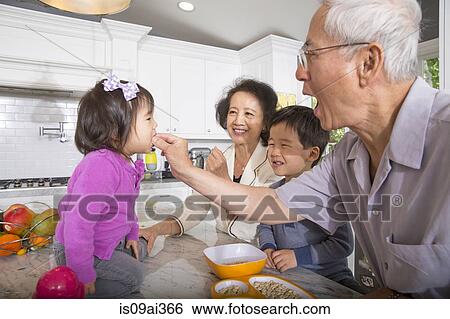 Stock Photograph - Grandfather feeding snack to toddler granddaughter in kitchen. Fotosearch