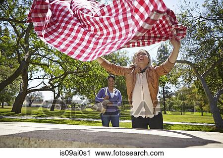 Stock Image - Lesbian couple preparing table cloth for picnic bench in park. Fotosearch