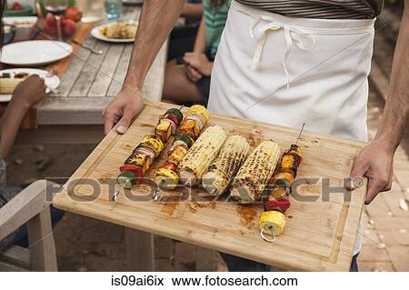 Stock Photo - Man holding tray of barbecued vegetable skewers and corn on the cob. Fotosearch