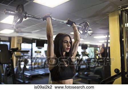 Stock Image - Mid adult woman using barbell in gym. Fotosearch
