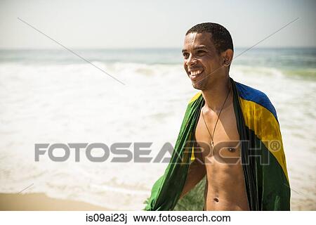 Stock Image - Portrait of young man wrapped in Brazilian flag, Arpoador beach, Rio De Janeiro, Brazil. Fotosearch