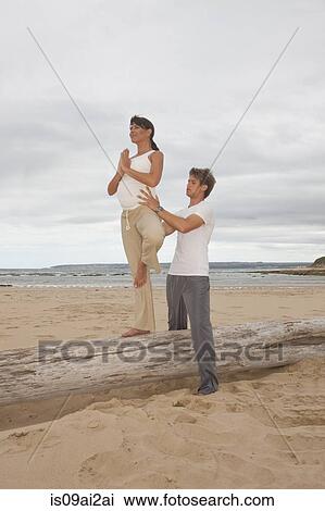 Stock Image - Pregnant mid adult woman with personal trainer practicing yoga at beach. Fotosearch