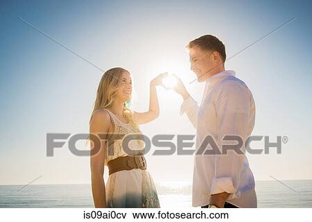 Stock Photo - Young couple making a heart in front of sun with hands. Fotosearch