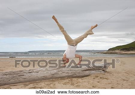 Stock Image - Young man doing handstand on tree trunk at beach. Fotosearch