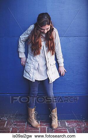 Stock Photo - Young woman leaning against blue wall looking down. Fotosearch