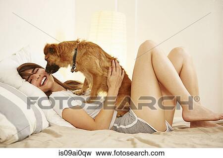 Stock Photo - Young woman lying on bed having face licked by pet dog. Fotosearch