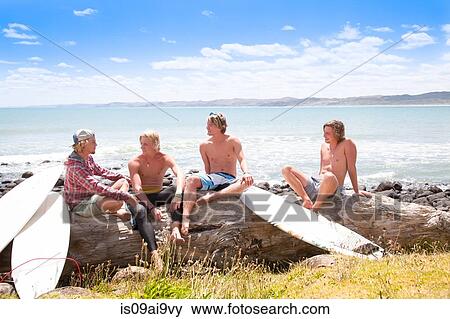 Stock Photo - Four young male surfer friends chatting on beach rocks. Fotosearch