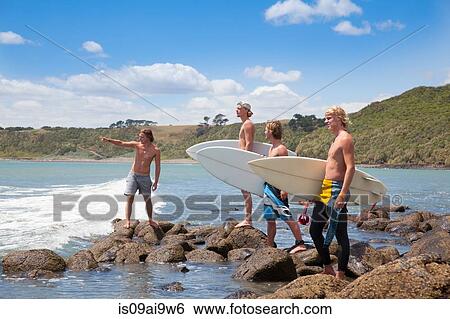 Stock Photograph - Four young male surfer friends pointing to sea from rocks. Fotosearch