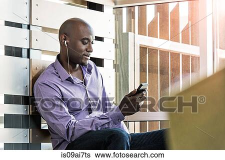 Stock Photograph - Male office worker listening to smartphone earphones in office. Fotosearch