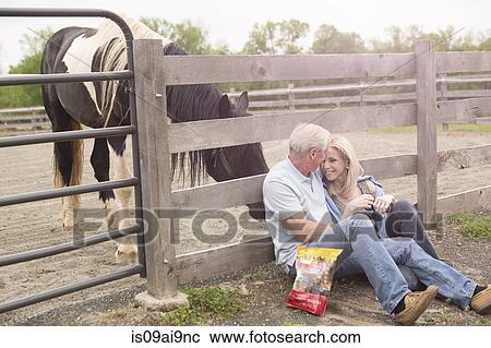 Stock Image - Mature couple sitting against fence and a curious horse. Fotosearch