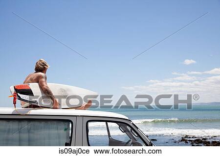 Stock Image - Mature male surfer watching from vehicle roof at beach. Fotosearch