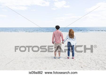 Stock Image - Rear view of couple holding hands on beach. Fotosearch