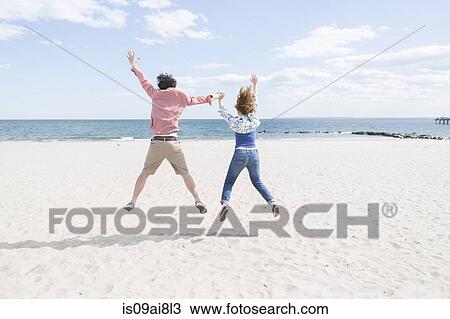 Stock Image - Rear view of couple jumping mid air on beach. Fotosearch