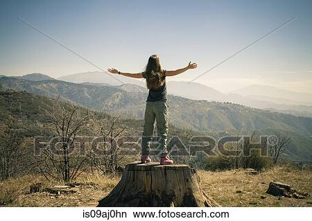 Picture - Rear view of mid adult woman on tree trunk with open arms, Lake Arrowhead, California, USA. Fotosearch