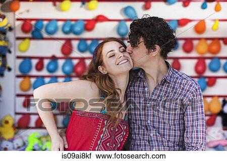 Stock Image - Romantic couple kissing on cheek at fairground stall. Fotosearch