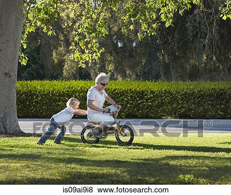 Stock Photo - Three year old boy pushing grandmother on cycle in park. Fotosearch