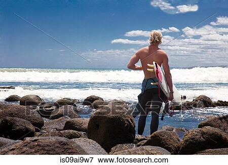 Stock Photo - Young adult male surfer watching sea from beach rocks. Fotosearch
