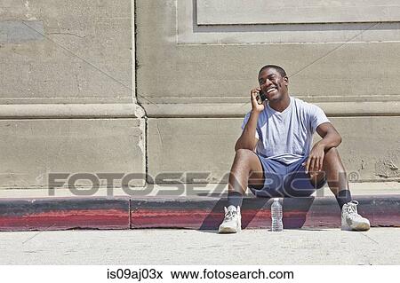 Stock Photo - Young male runner sitting on sidewalk chatting on smartphone. Fotosearch