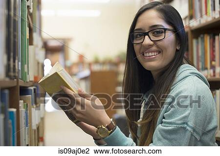 Stock Image - Young woman holding book in library. Fotosearch