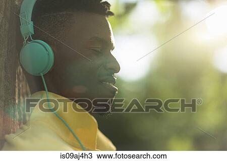 Stock Image - Close up of young man with eyes closed listening to headphones in park. Fotosearch