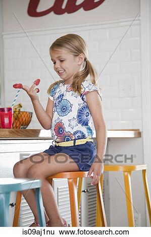 Stock Photograph - Girl sitting on kitchen stool with raspberries on her fingers. Fotosearch