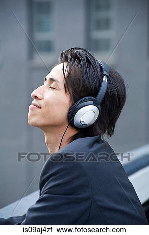 Stock Image - Mid adult businessman on city rooftop listening to music on headphones. Fotosearch