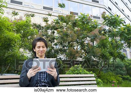 Picture - Mid adult businessman sitting on park bench looking at digital tablet. Fotosearch
