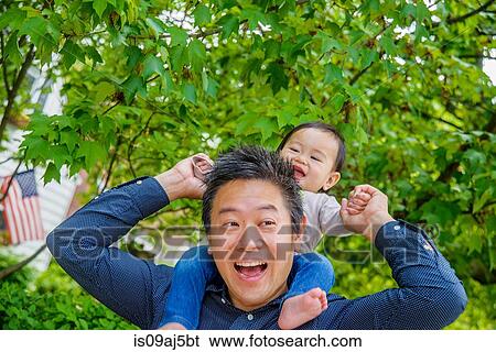 Stock Photograph - Mid adult father shoulder carrying baby son in garden. Fotosearch